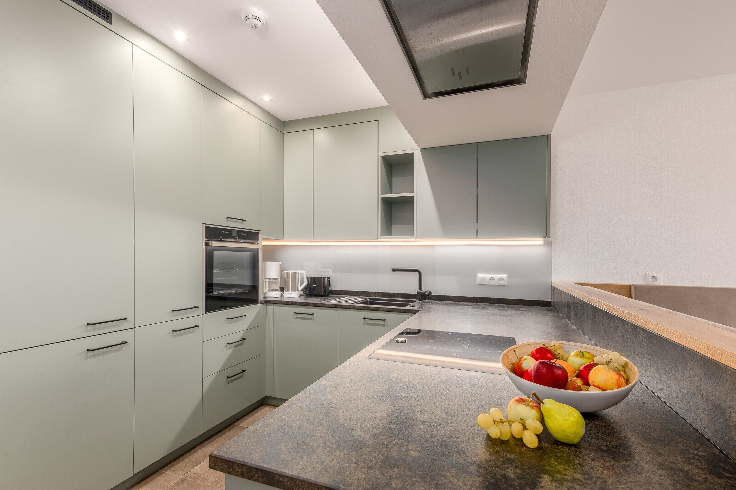 A modern kitchen with light green cabinets, a granite countertop, and a bowl of fruit.