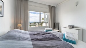 Ein Hotelzimmer mit Bett, Nachttisch, Lampe, einem Fenster mit Vorhängen, Schreibtisch und einer kleinen Schachtel auf dem Schreibtisch.