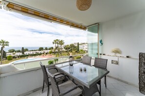 En balkong med ett glasbord och flätade stolar, med utsikt över en pool och grönområden.