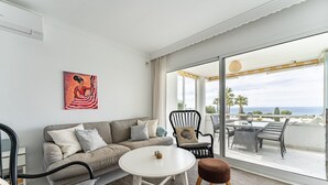 Ein modernes Wohnzimmer mit einer Couch, einem runden Tisch und einem Sessel. Ein großes Fenster bietet Blick auf das Meer.