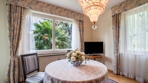 A room with a round table, a chair, a television, and a window with curtains. - Villa Coris Apartment Magnolia 110 sqm with Garden (Merano)