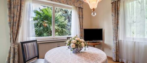 A room with a round table, a chair, a television, and a window with curtains.