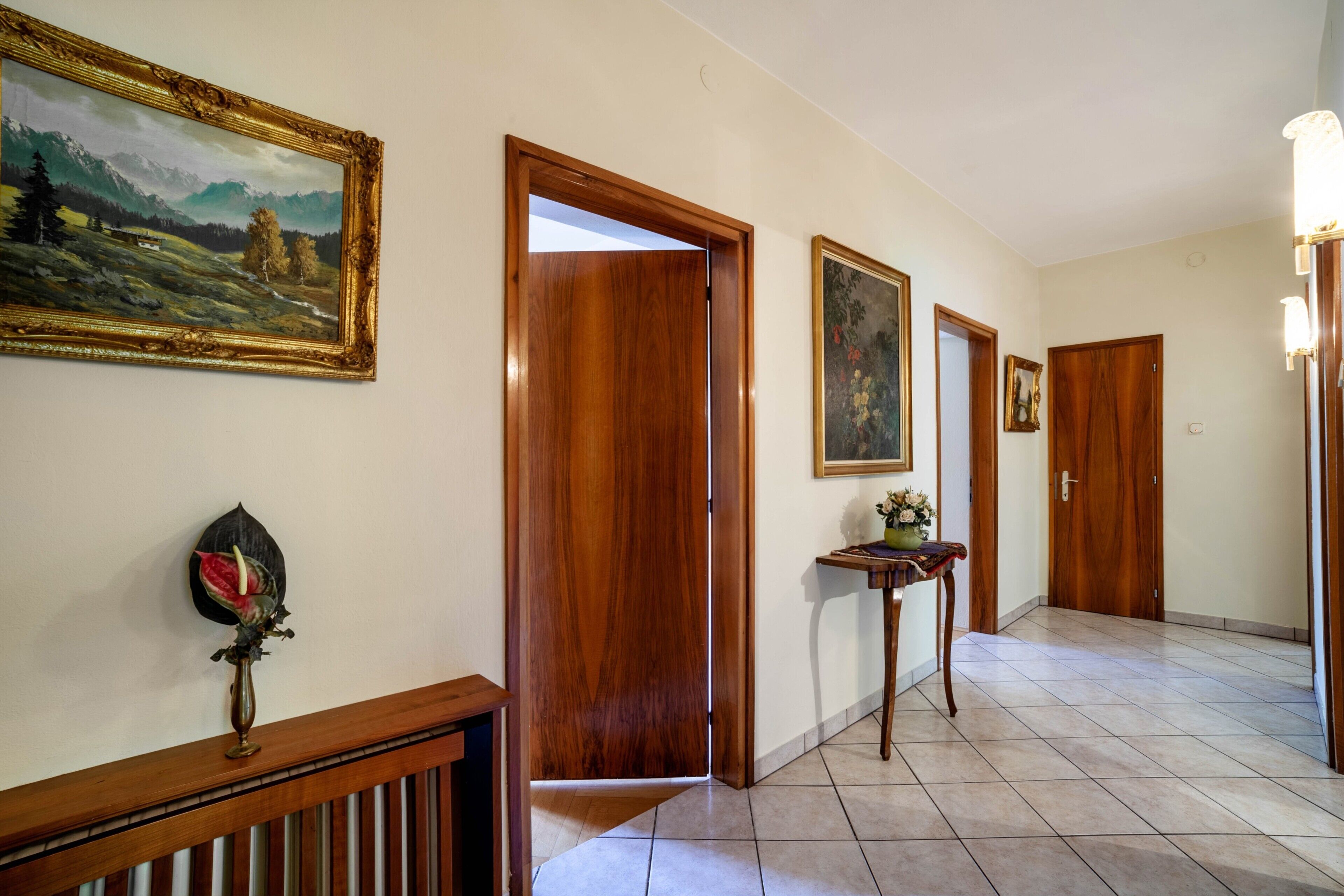A hallway with wooden doors, a framed landscape painting, a small table with a vase, and a staircase with a wooden railing.