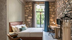 A bedroom with a stone wall, a bed with a wooden headboard, a desk with a chair, and a hanging garment. - Cohort Koukaki (Athens)