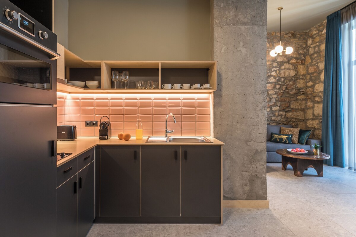 A modern kitchen with a built-in oven, a countertop with a toaster, and a sink. There is a stone wall and a sofa in the adjacent living area.