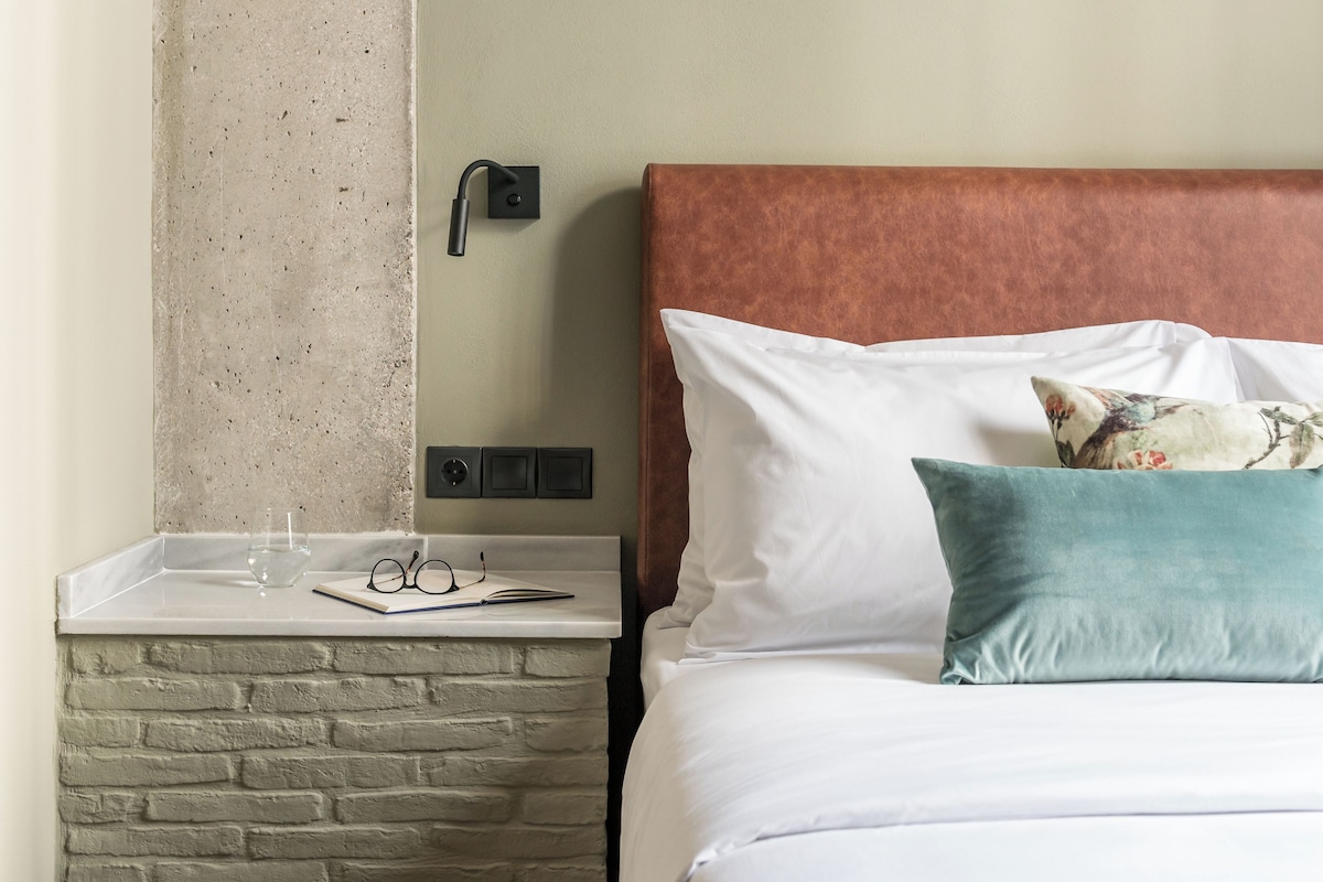 Una cama con ropa blanca y un cabecero marrón, una mesita de noche con textura similar a la piedra, un vaso de agua y un par de gafas.