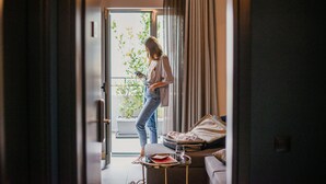 A woman standing by a sliding glass door in a room with a suitcase, a small table, and a sofa. - Cohort Koukaki (Athens)