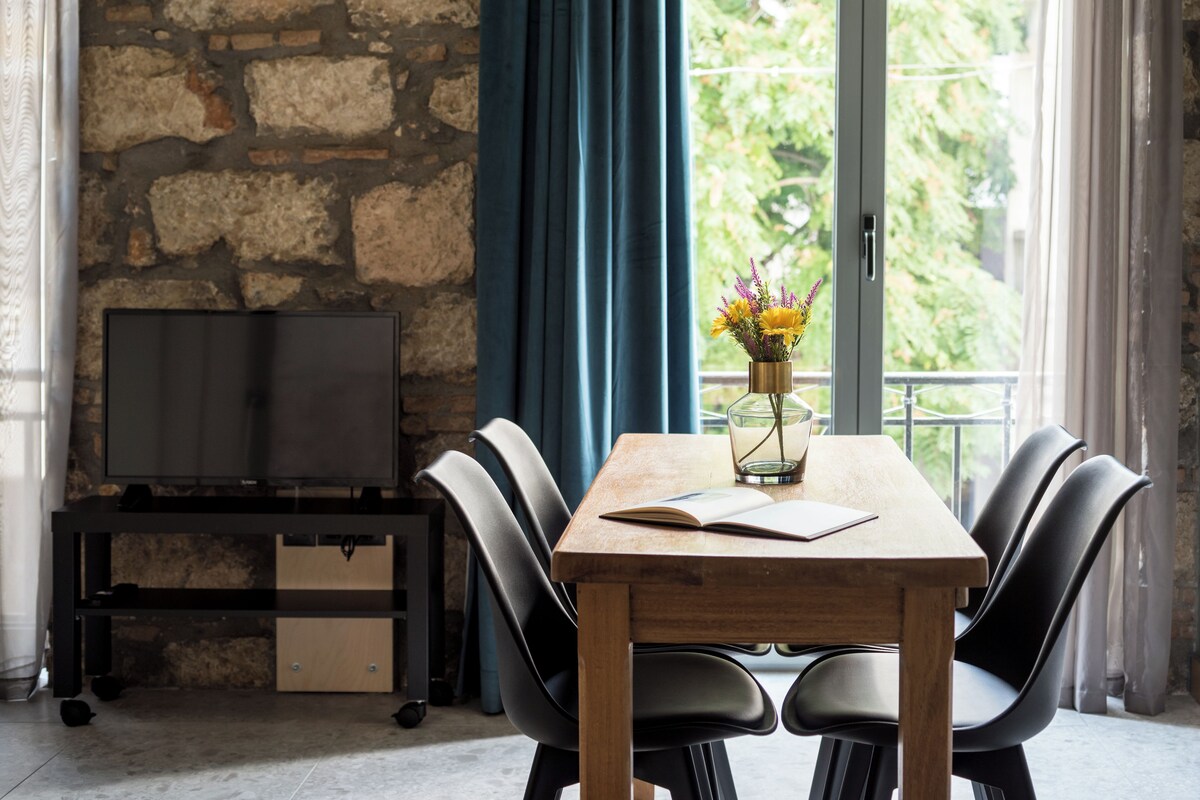 Un coin repas avec une table en bois, des chaises noires, un téléviseur posé sur un meuble et une vue sur la verdure grâce à de grandes fenêtres.