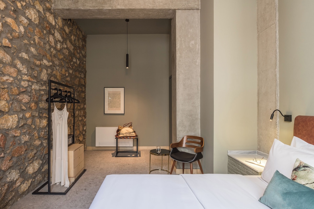 Habitación de hotel con pared de piedra, cama con ropa de cama blanca, silla de madera, mesita con vaso y lámpara colgante.
