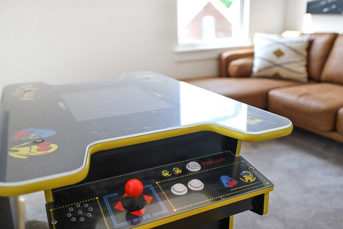 Una mesa de juegos arcade pequeña con un joystick y botones, ubicada en una sala de estar con un sofá de cuero marrón y un cuadro en la pared.
