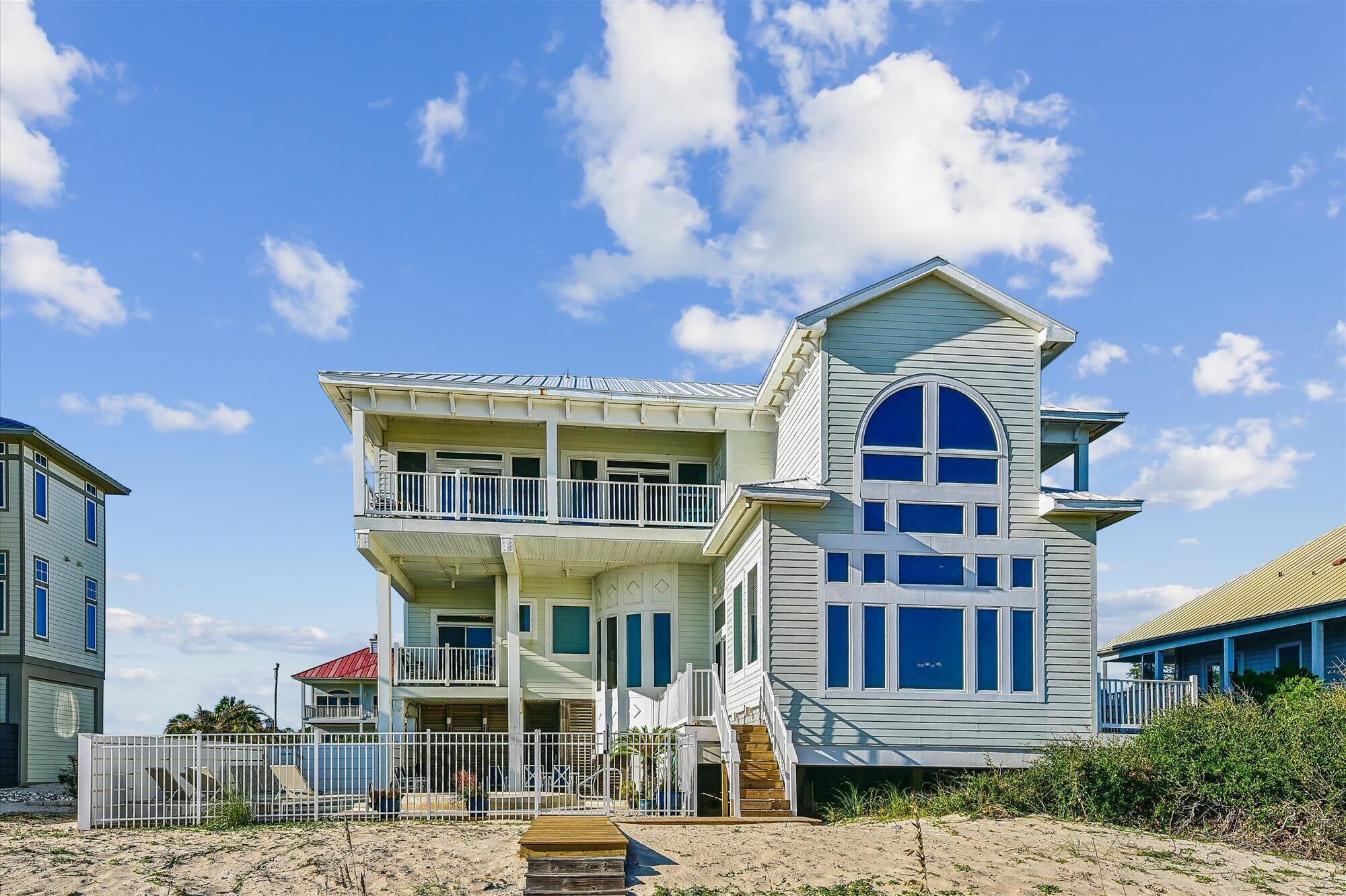 A two-story beachfront house with a balcony, a large arched window, and a staircase leading to the upper floor.
