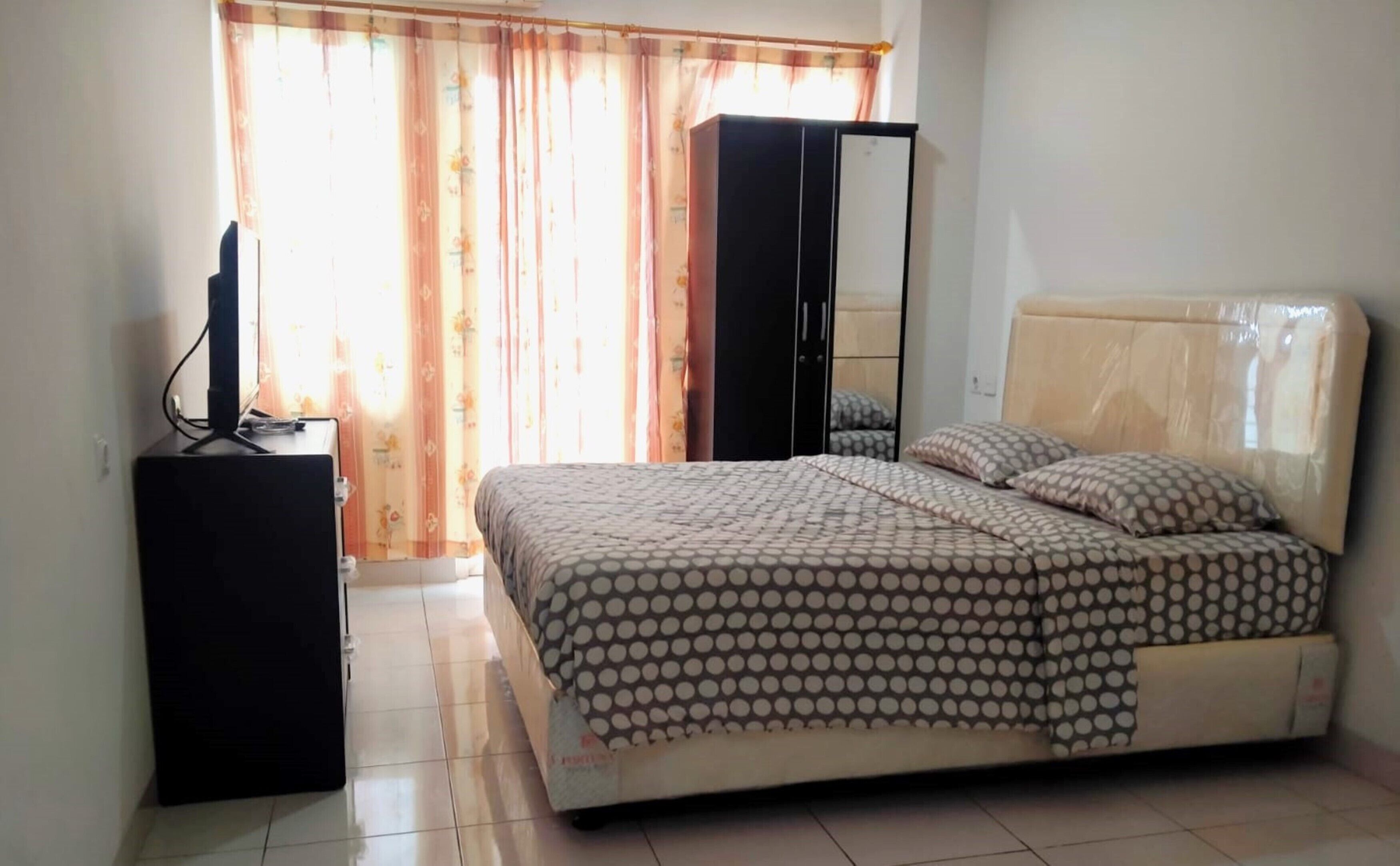 A bedroom with a bed, a television on a stand, a wardrobe, and a window with curtains.