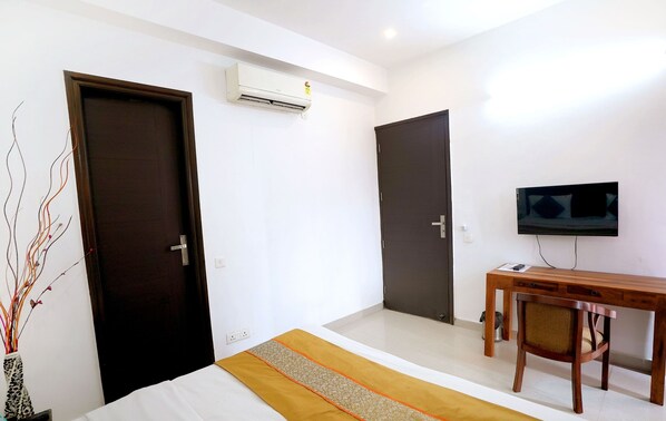 A hotel room with two doors, a bed with a striped bedspread, a wooden desk with a chair, and a flat-screen TV mounted on the wall.
