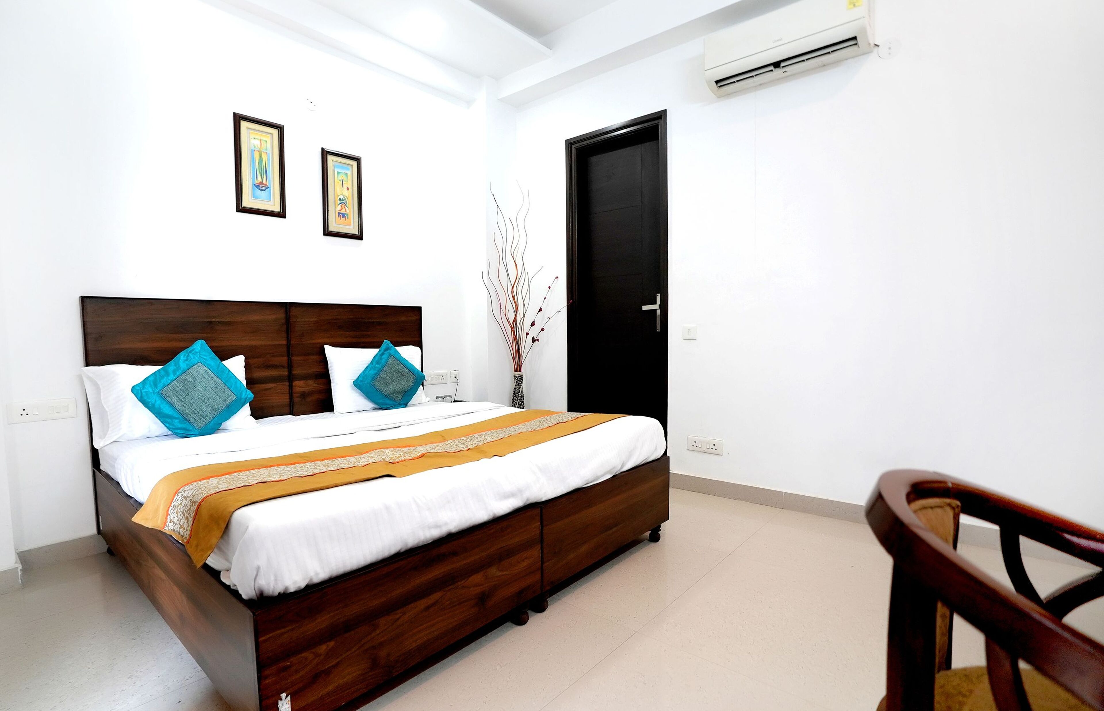 A neatly made bed with a wooden headboard, white bedding, and decorative pillows. There is a black door and an air conditioning unit on the wall.