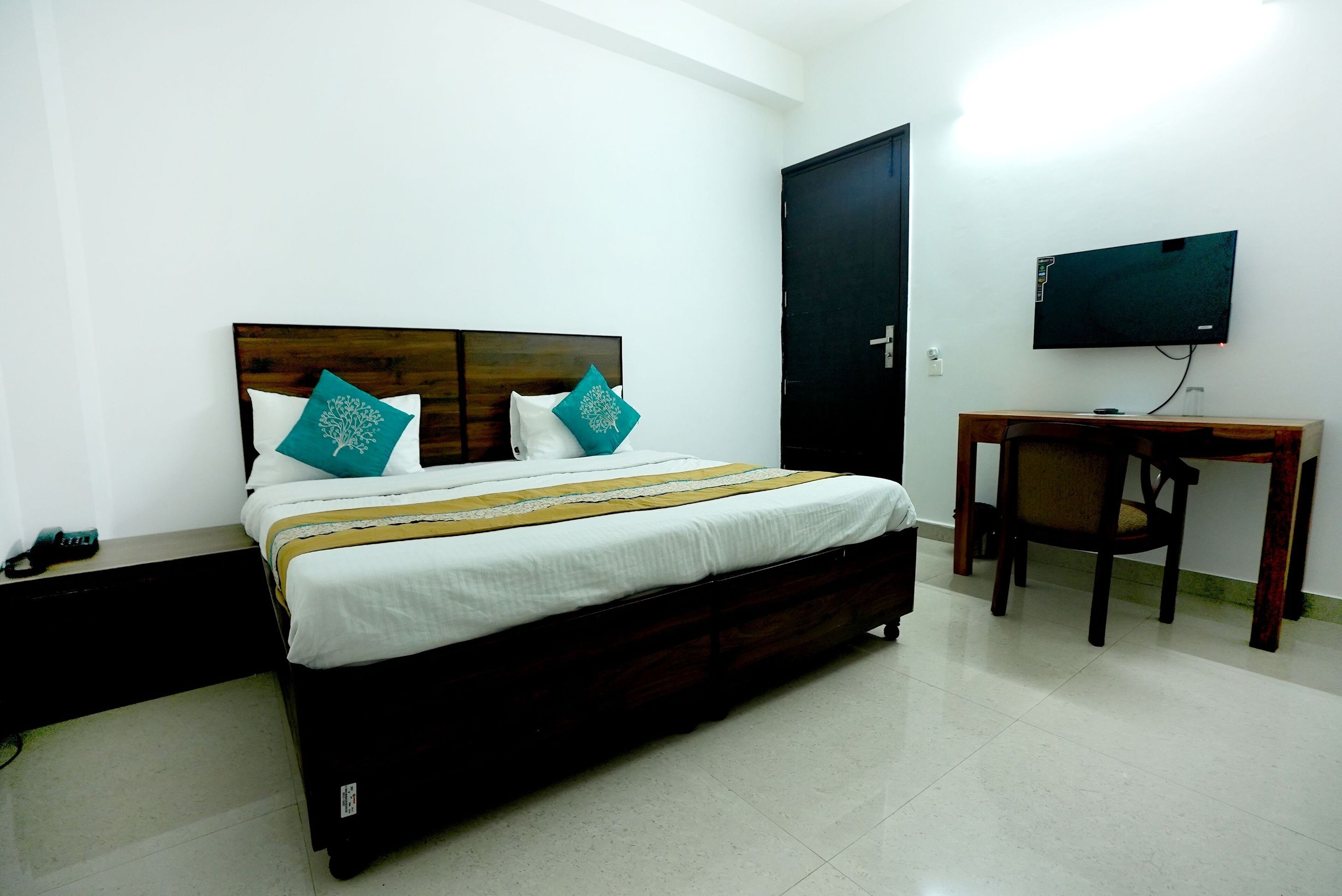 A bedroom with a wooden bed, white bedding, teal pillows, a desk with a chair, and a TV on the wall.