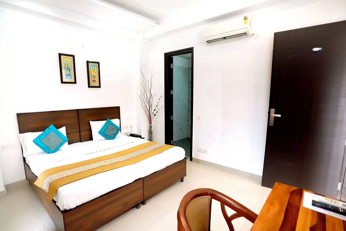 A hotel room with a wooden bed, a desk, and a chair. There is a door, an air conditioning unit, and two framed pictures on the wall.
