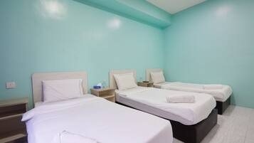 Habitación de hotel con tres camas, cada una con ropa de cama y almohadas blancas. Hay mesitas de noche de madera con lámparas y una pequeña mesa entre las camas. Las paredes están pintadas de color verde azulado y el piso está cubierto de baldosas.
