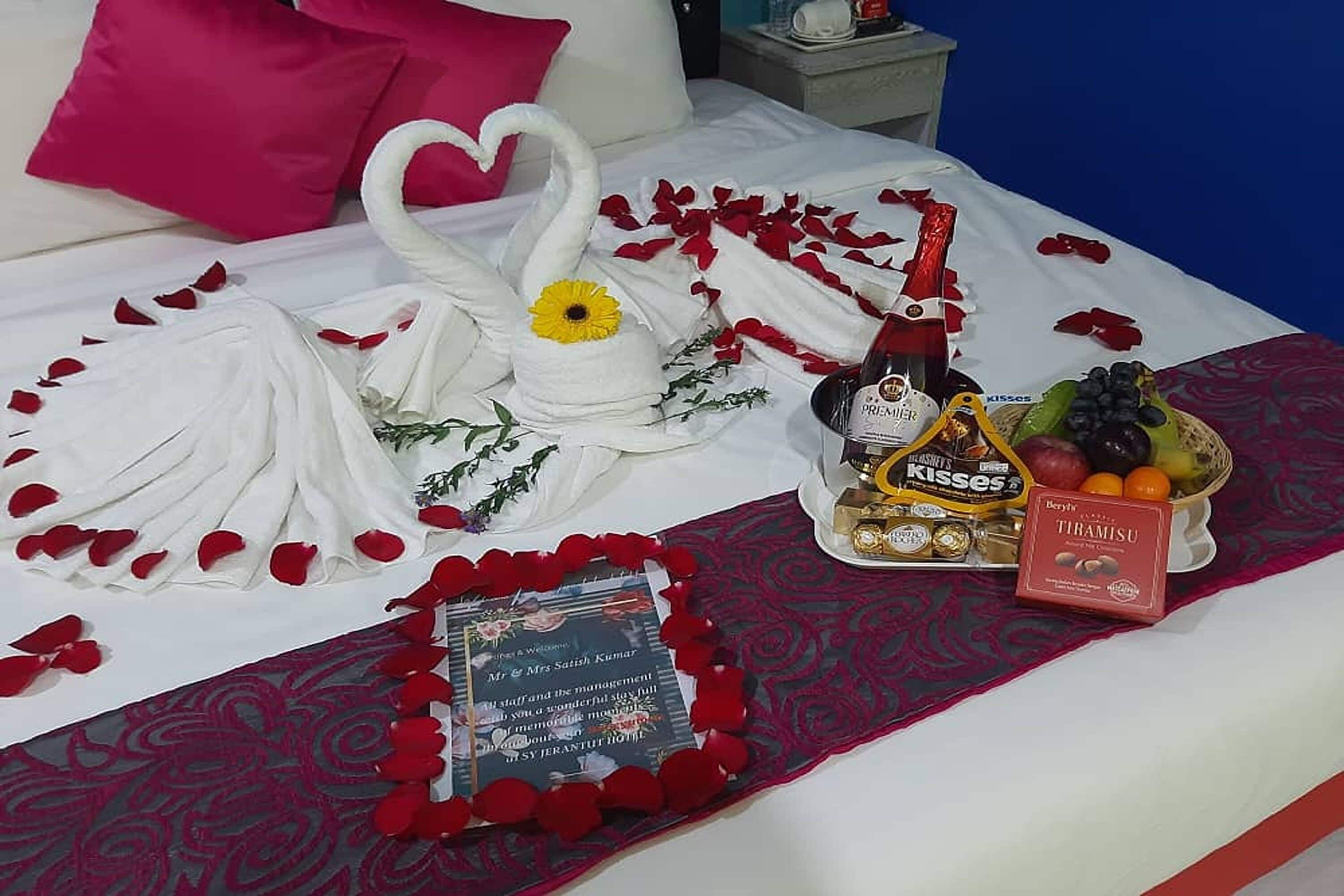 Uma cama com pétalas de rosa, uma toalha em formato de cisne, um buquê de flores, uma garrafa de champanhe, uma caixa de chocolates e uma cesta de frutas.