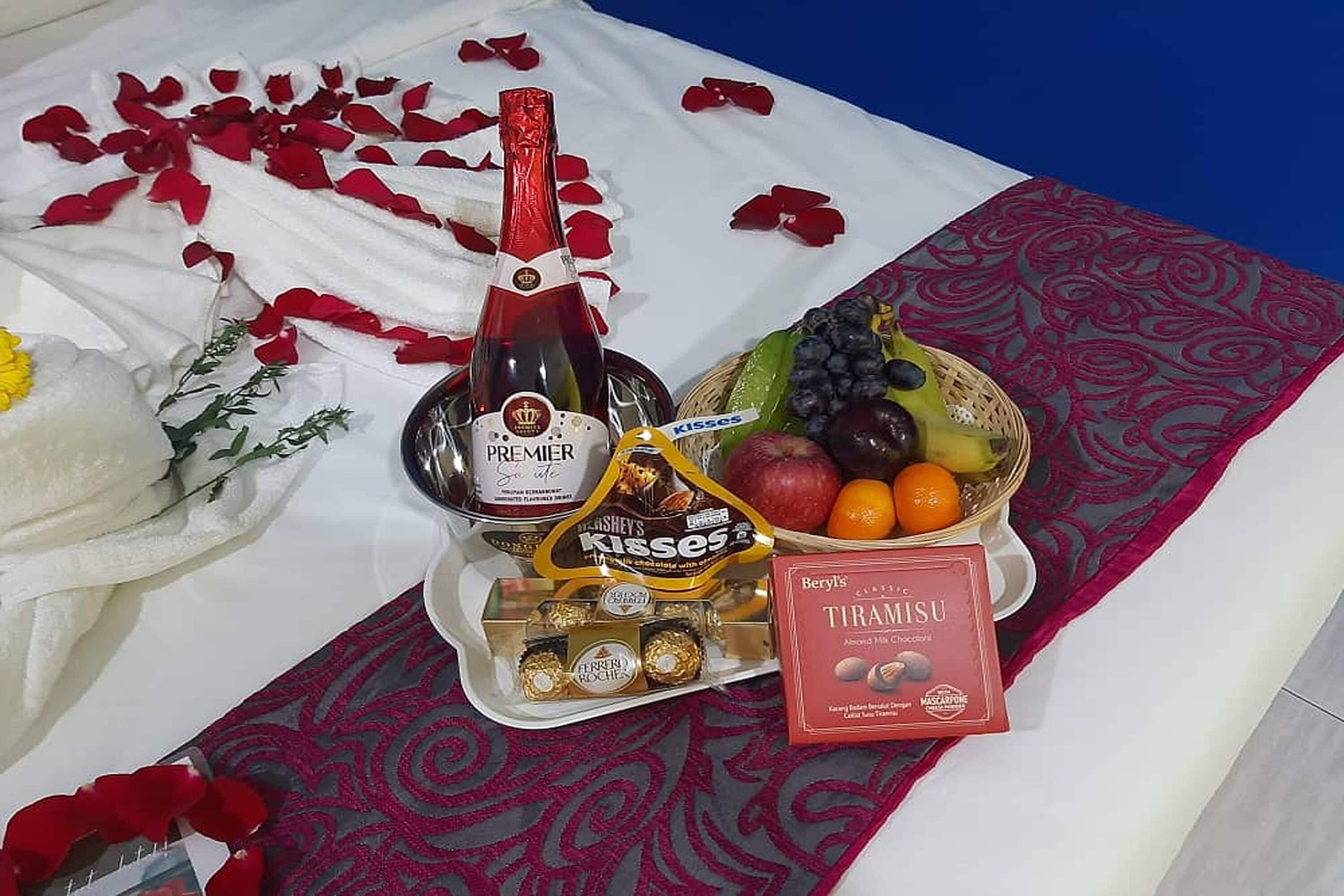 Una cama con una manta blanca y roja, una botella de champán, un cuenco de fruta, bombones y una caja de galletas.