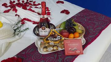 Una cama con una manta blanca y roja, una botella de champán, un cuenco de fruta, bombones y una caja de galletas.