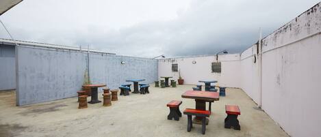 An outdoor patio area with multiple tables and benches, surrounded by walls and a cloudy sky.