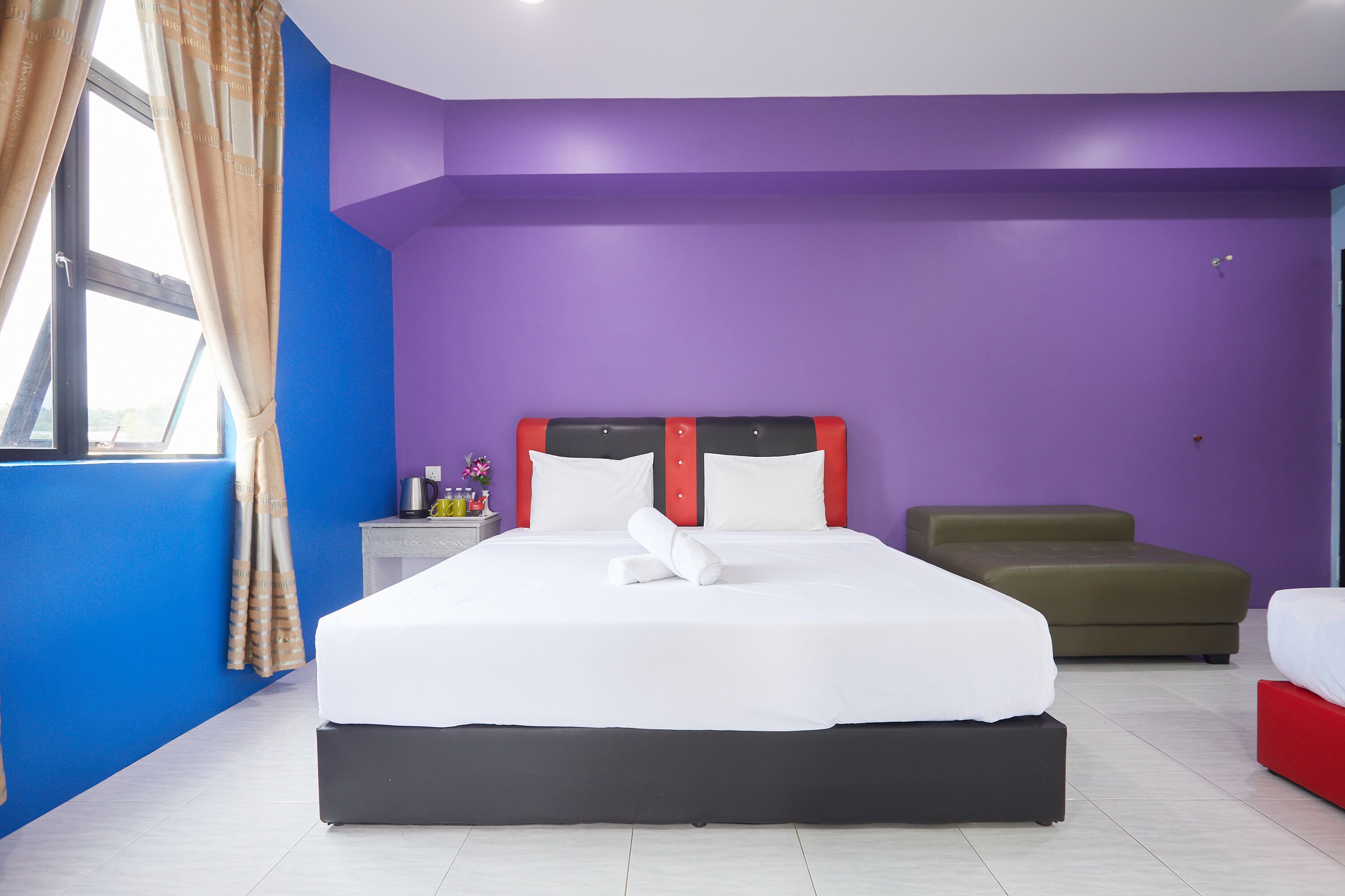 A hotel room with a large bed, a nightstand, a green ottoman, and a window with curtains.