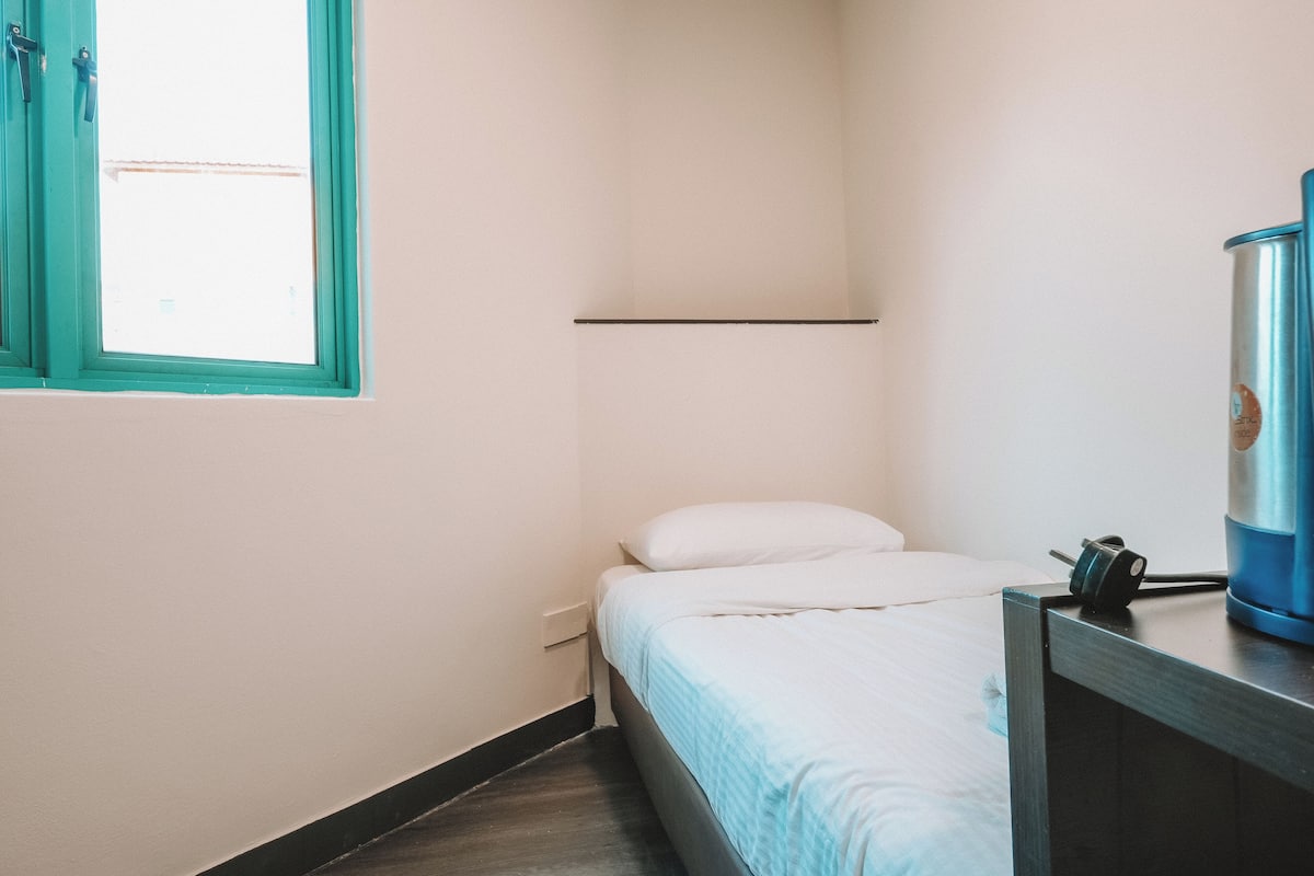 Una habitación pequeña y estrecha con una cama individual, un ventanal con detalles color turquesa y una botella de agua azul sobre una mesita de noche de madera.