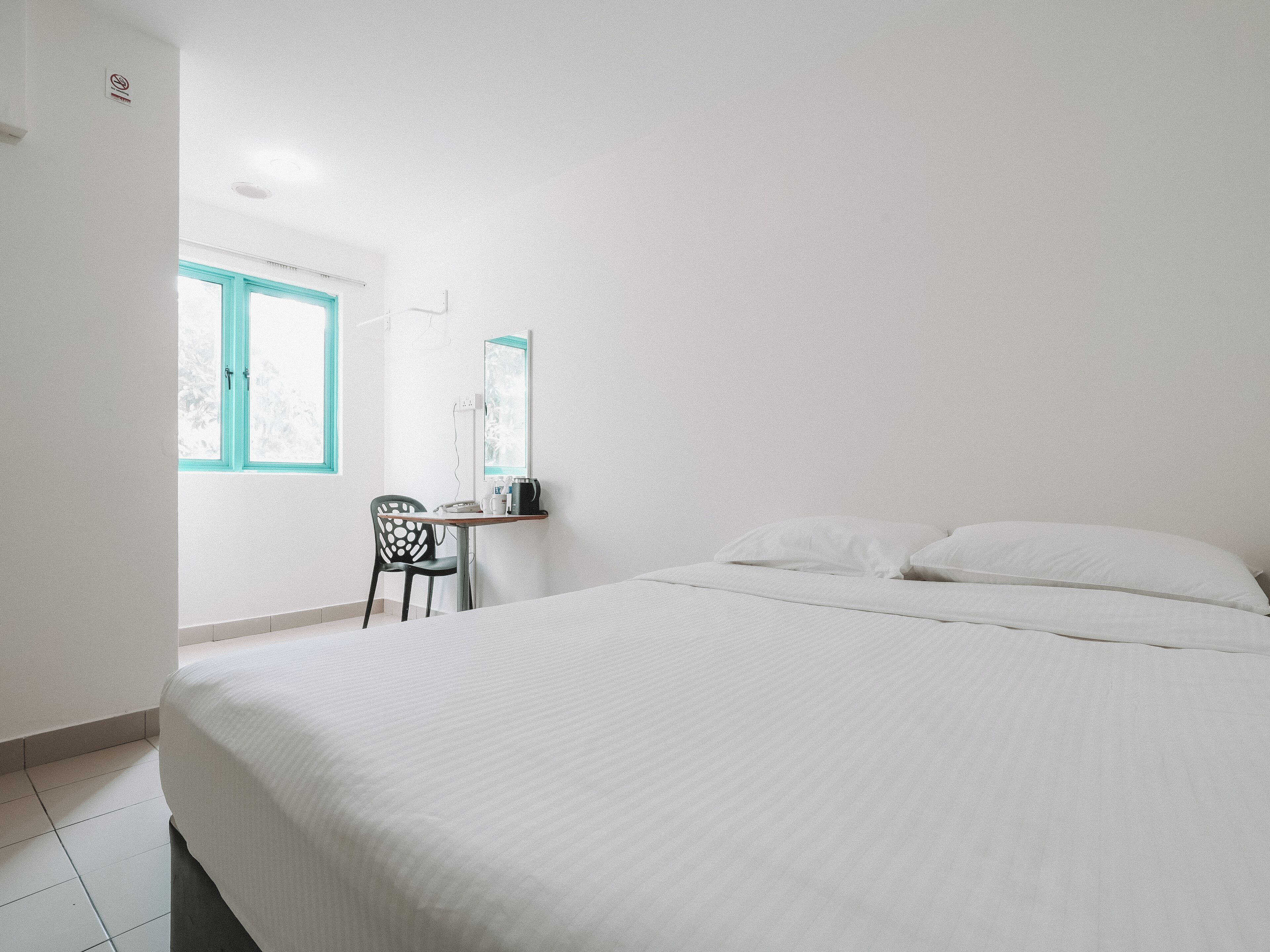 Quarto de hotel minimalista com uma cama, uma escrivaninha com cadeira, uma janela e um espelho fixado na parede.