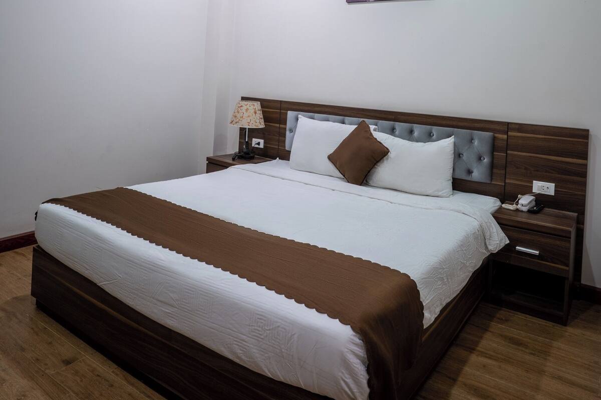 Un lit bien fait, recouvert d’une couverture marron et de draps blancs, une table de chevet avec une lampe et un tableau accroché au mur.