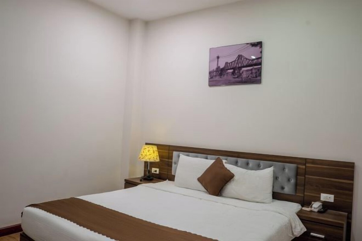 Une chambre d’hôtel avec un lit, une table de chevet avec une lampe, un téléphone et un tableau représentant un pont, accroché au mur.