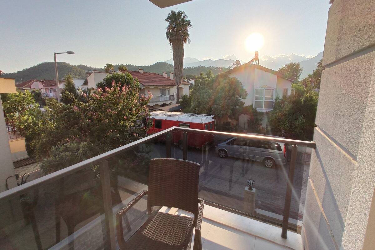 Un balcón con una silla de mimbre, vista a una calle con un camión de bomberos rojo y una montaña al fondo.