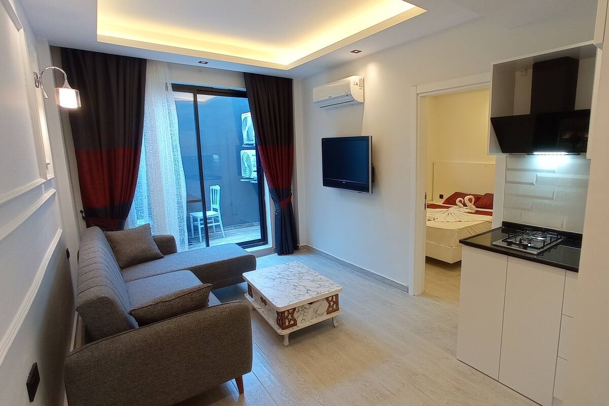 Una sala moderna con un sofá, una mesa de centro y un televisor. Hay un ventanal corredero que da a un balcón.