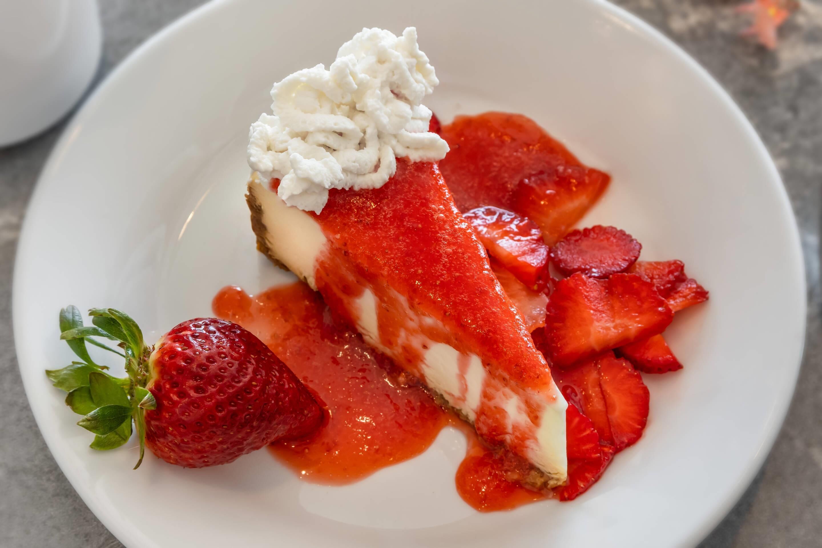 A dessert plate with a slice of cheesecake topped with whipped cream and strawberry sauce, accompanied by fresh strawberries.