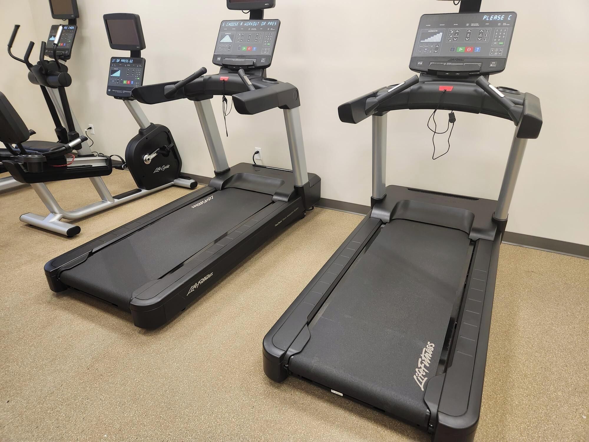 Dos cintas de correr con pantallas digitales y paneles de control, una bicicleta estática y un banco de pesas en un gimnasio.