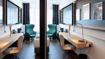 A modern hotel room with a mirrored wall, a wooden desk, two teal chairs, a bed, and artwork on the walls.