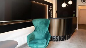 A teal chair and a small table in a modern lobby with a reception desk and a sign that reads "DESIGN". - Design- und Kunsthotel München (Munich)
