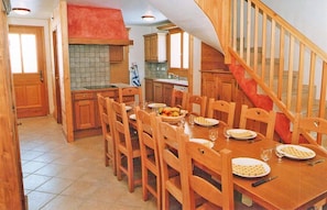 Un comedor con una mesa de madera dispuesta para cuatro personas, rodeada de sillas de madera. Se ve una cocina con armarios de madera y una campana extractora roja al fondo.