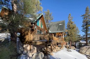 A log cabin with a wooden deck and stone foundation, surrounded by pine trees and snow.