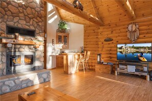 Interior acogedor de una cabaña con chimenea de piedra, muebles de madera y un televisor que muestra una vista panorámica.
