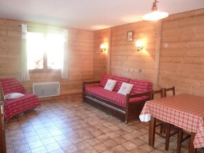 Une pièce chaleureuse avec un plancher en bois, un canapé rouge agrémenté de coussins, une petite table recouverte d’une nappe à carreaux et une fenêtre avec des rideaux.