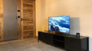 A modern living room with a flat-screen TV on a black entertainment unit, a wooden door, and a tiled floor. - Wifi, Ground floor, view mountain, terrace, parking, ski locker, 40m², Le Grand Bornand (Le Grand-Bornand)