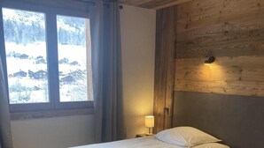 A room with two beds, wooden flooring, a window with a view of snow-covered mountains, and a wall-mounted light fixture. - Wifi, 50m from ski slopes, Ground floor, view mountain, terrace, fireplace or stove, parking, 55m² (Le Grand-Bornand)