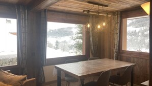 A wooden dining area with a table and chairs, large windows with curtains, and a view of a snowy landscape. - Wifi, 50m from ski slopes, Ground floor, view mountain, terrace, fireplace or stove, parking, 55m² (Le Grand-Bornand)