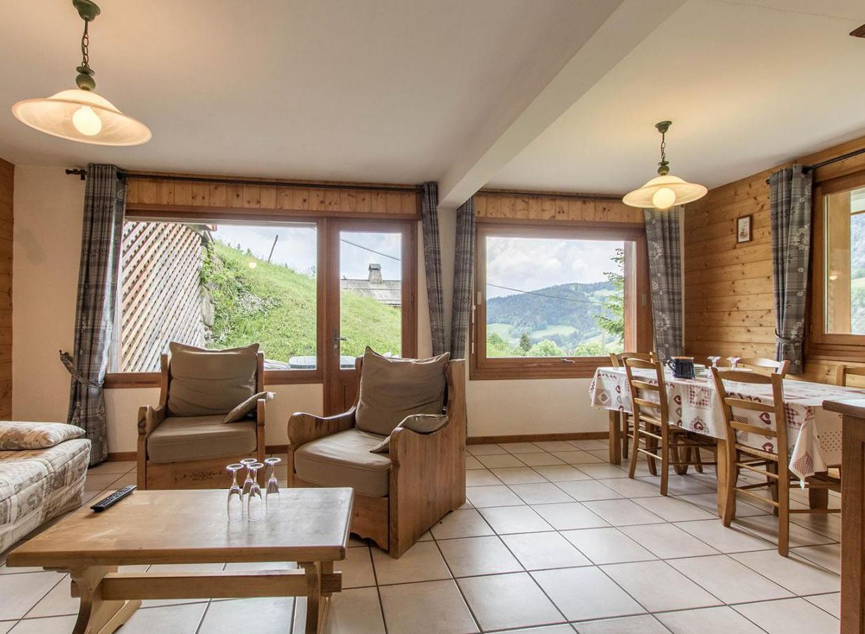 A cozy room with a sofa, two armchairs, a dining table, and chairs. Large windows offer a view of the outdoors.