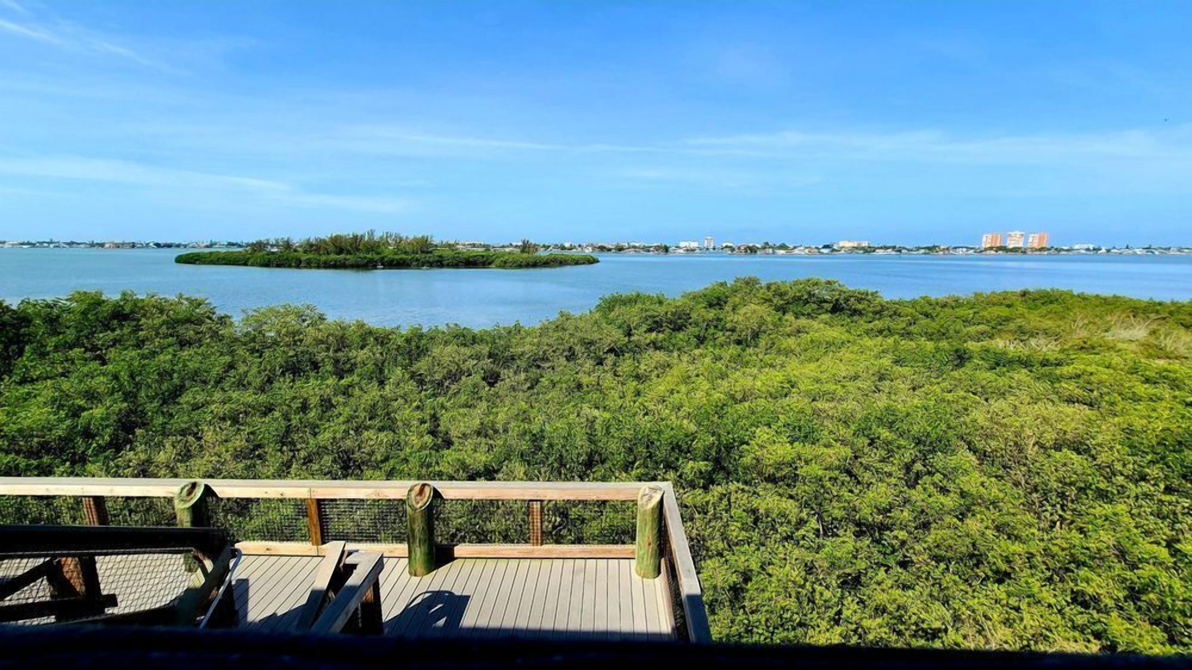 Blick von einer Holzterrasse auf ein Gewässer mit einer fernen Insel und Gebäuden.