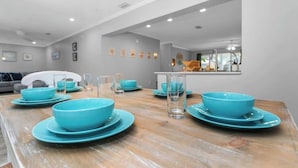 Un comedor con una mesa de madera dispuesta con platos y tazones azules, un sofá blanco al fondo y una cocina visible a través de un arco.
