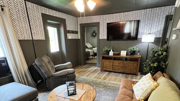 Ein modernes Wohnzimmer mit Sofa, Sessel und Couchtisch. Ein Fernseher ist an der Wand befestigt und ein Deckenventilator vorhanden.
