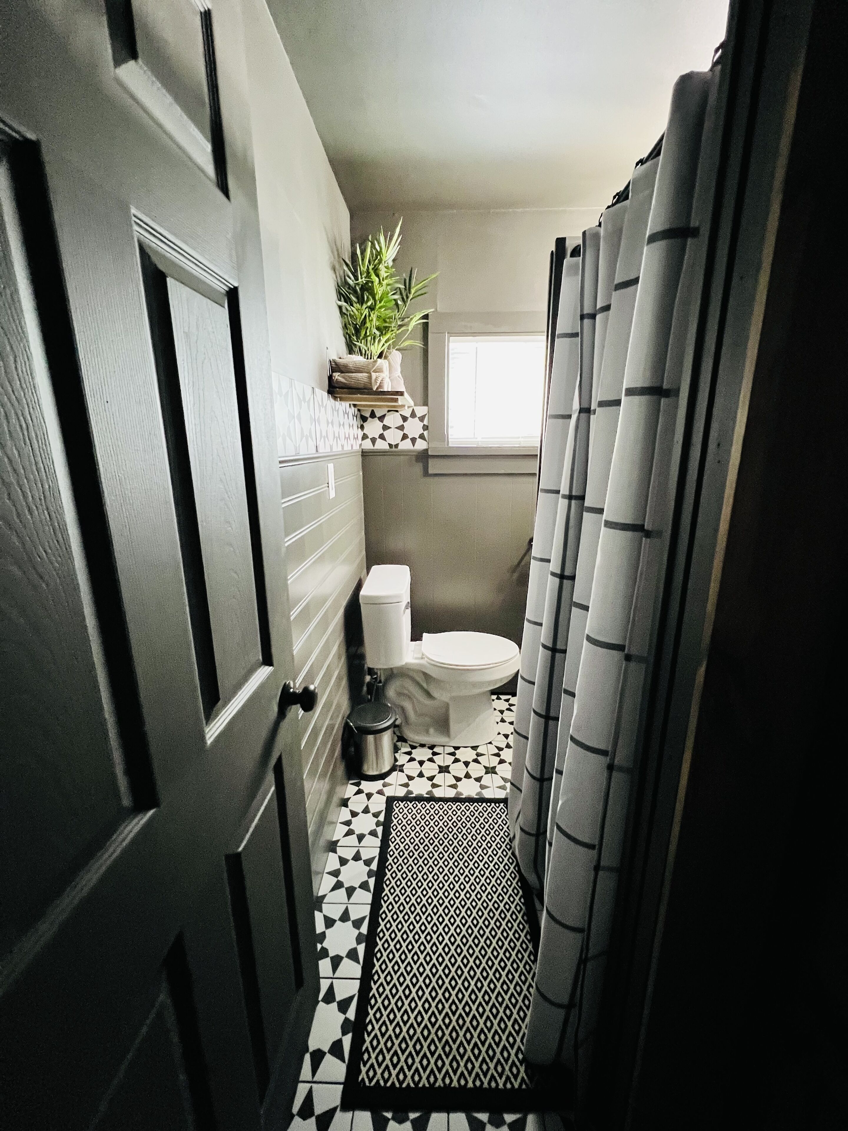 Een badkamer met een zwart-wit betegelde vloer, een toilet, een prullenbak, een plant op een plank en een douchegordijn.