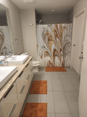 Une salle de bain moderne avec un grand miroir, deux lavabos, des toilettes et une douche agrémentée d’un rideau décoratif.