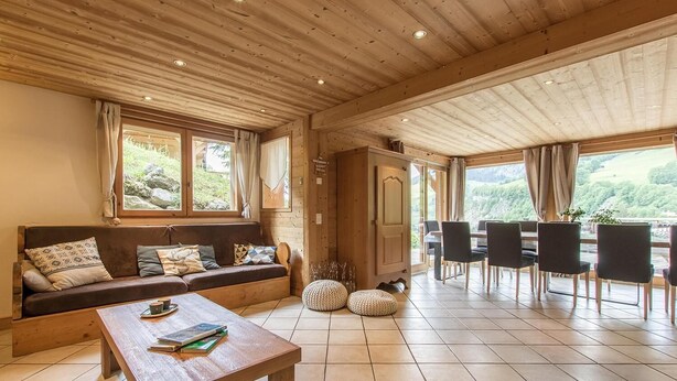 A cozy room with a wooden ceiling, a sofa with cushions, a dining table with chairs, and large windows offering a view of greenery.